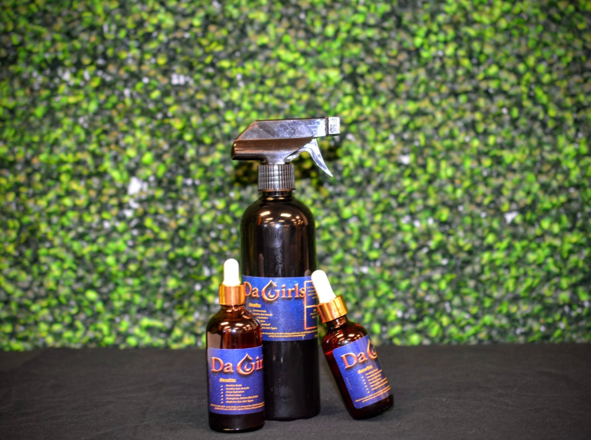 Set of 'Da Girls' hair care products including a spray bottle and two bottles with droppers against a green textured wall.